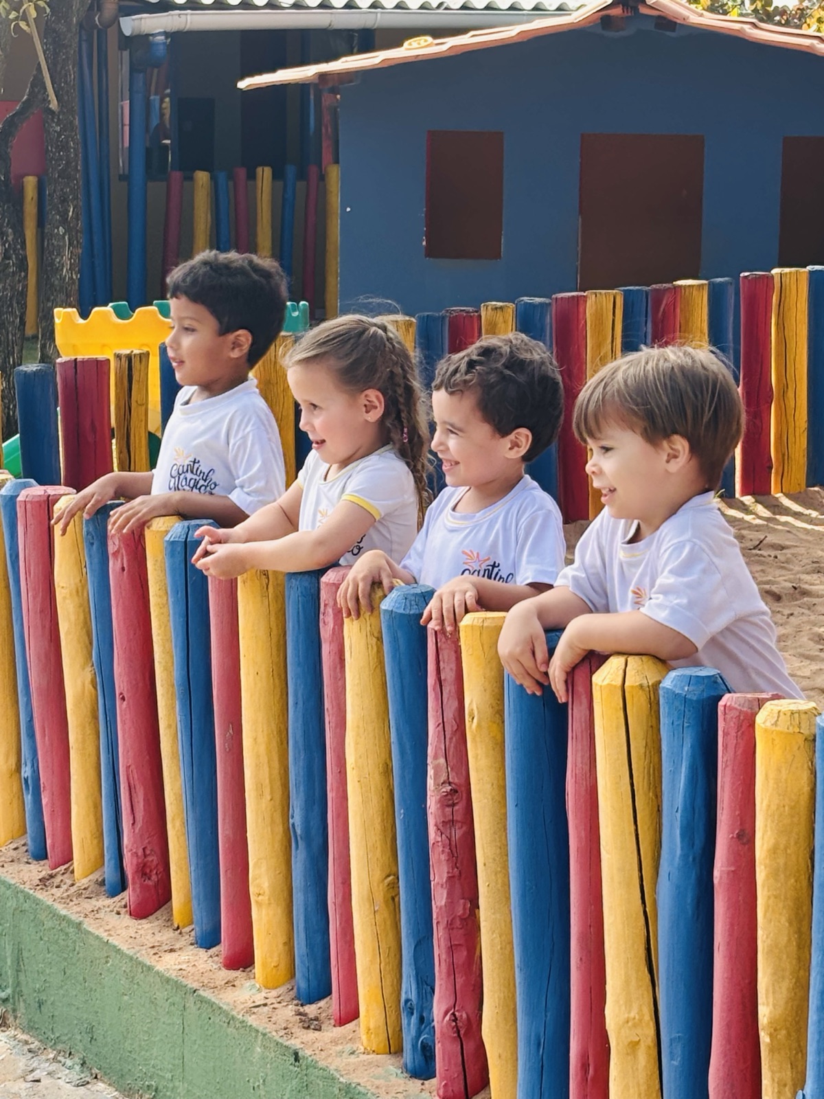 Crianças pequenas sorrindo apoiadas na cerca colorida do parquinho do Cantinho Mágico